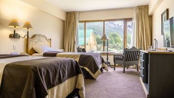 A hotel room with two beds, a desk, a chair, and a large window offering a mountain view.