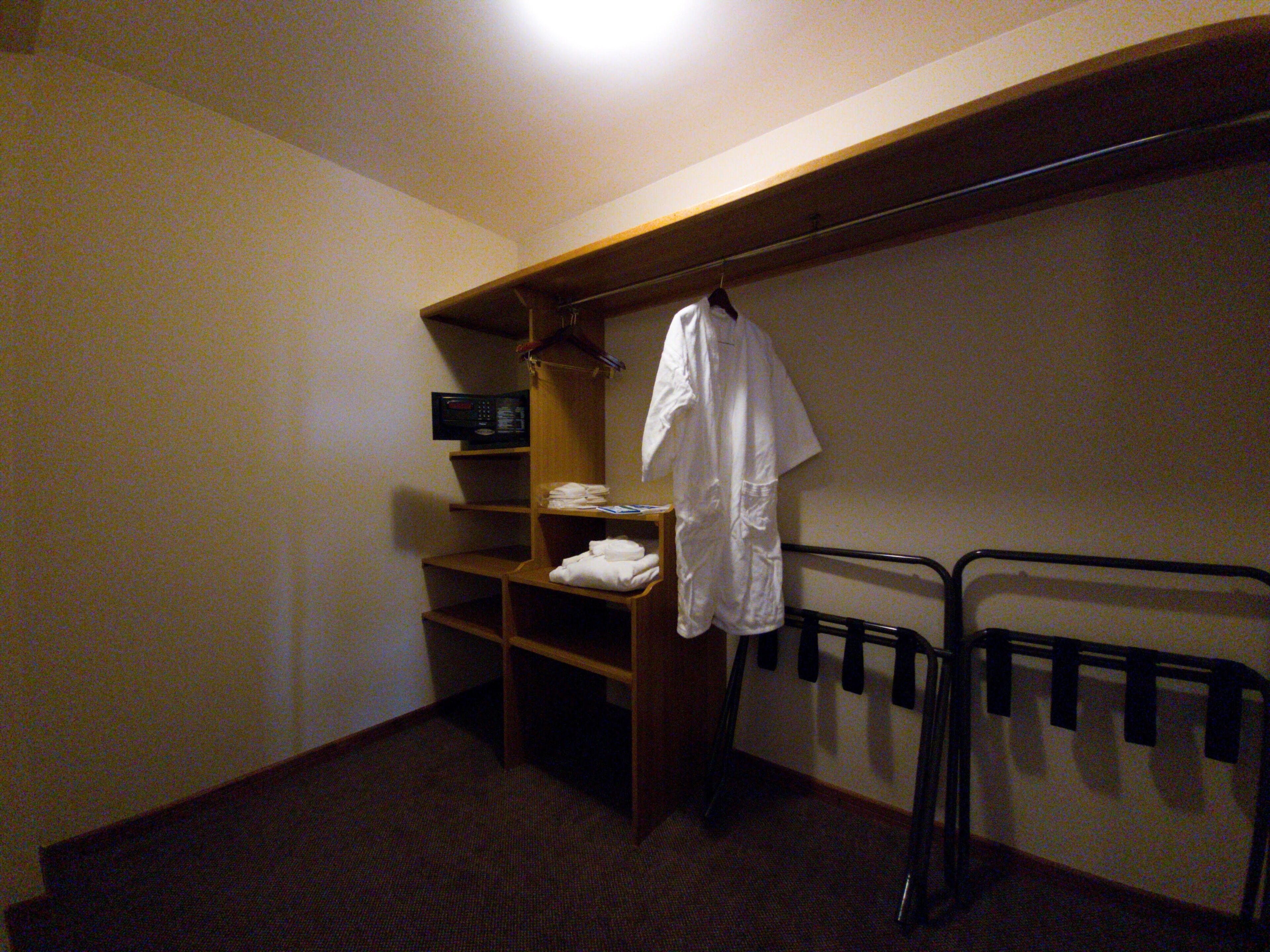 Una habitación pequeña, con poca luz, que tiene un estante de madera con una camisa blanca, una toalla doblada y un pequeño dispositivo electrónico. En la pared, hay un perchero metálico con ganchos.
