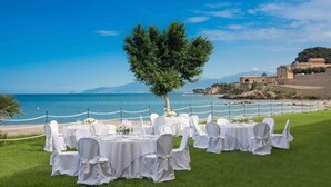 An outdoor wedding setup with round tables and chairs, a tree, and a coastal backdrop. - Tonnara di Trabia (Trabia)