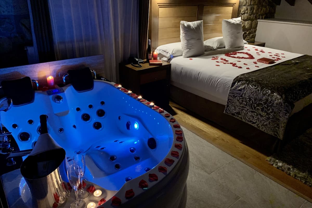 Een slaapkamer met een stenen muur, een houten bed met rozenblaadjes, een nachtkastje met telefoon en kaars, en een bubbelbad met rozenblaadjes.