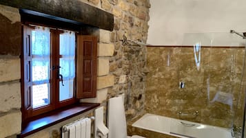 Un baño con pared de piedra, ventana de madera, inodoro blanco, ducha con mampara de vidrio y lavamanos blanco.