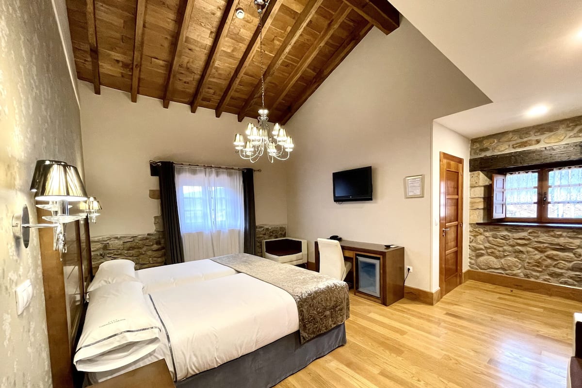Een slaapkamer met een houten plafond, een bed met een witte dekbedovertrek, een bureau met een computer, een televisie aan de muur, en een stenen muur met ramen.