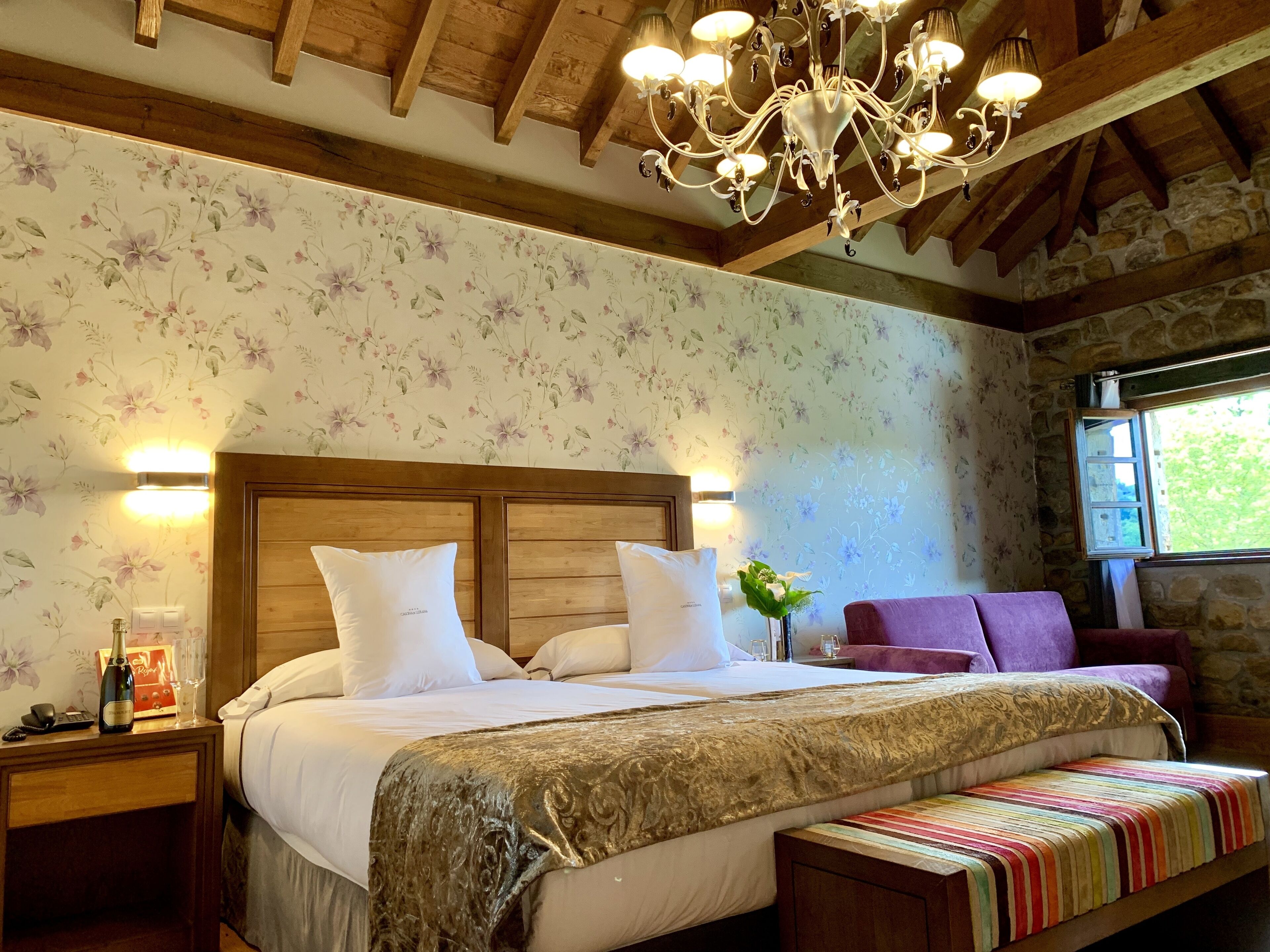 A bedroom with a wooden headboard, a large bed with white bedding, a colorful striped bench, a purple armchair, a chandelier, and a floral wallpaper.