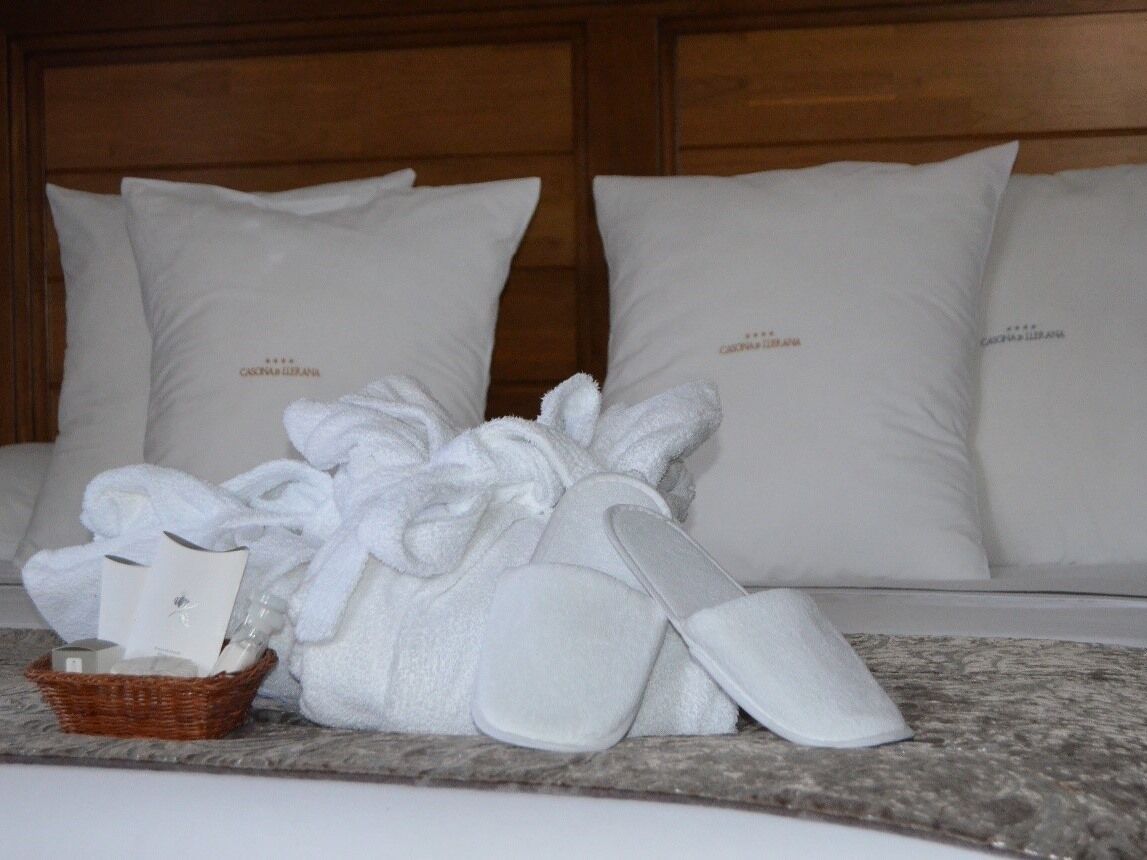 Quarto de hotel com cama, almofadas brancas, roupão, chinelos e um cesto com produtos de higiene pessoal.
