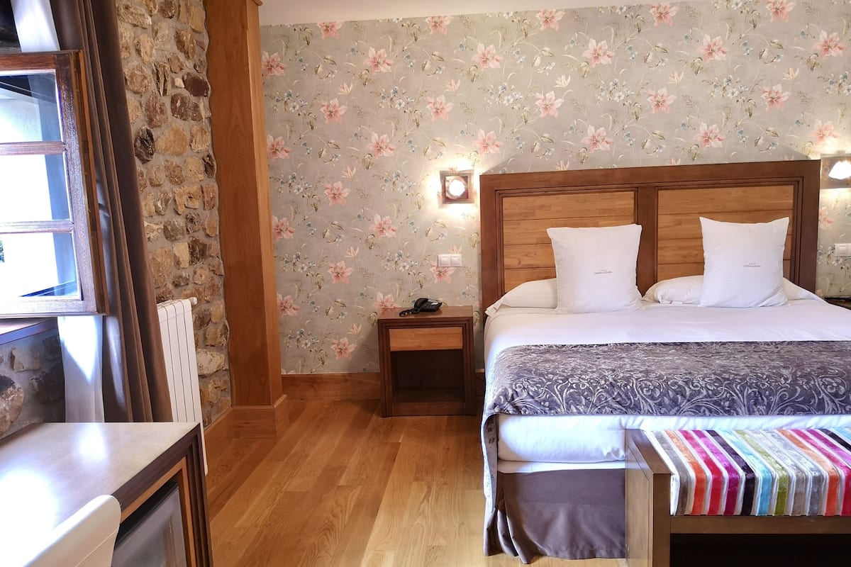 Een slaapkamer met een houten hoofdbord, een bed met witte beddengoed, een nachtkastje met een lamp, een stenen muur en een raam met gordijnen.