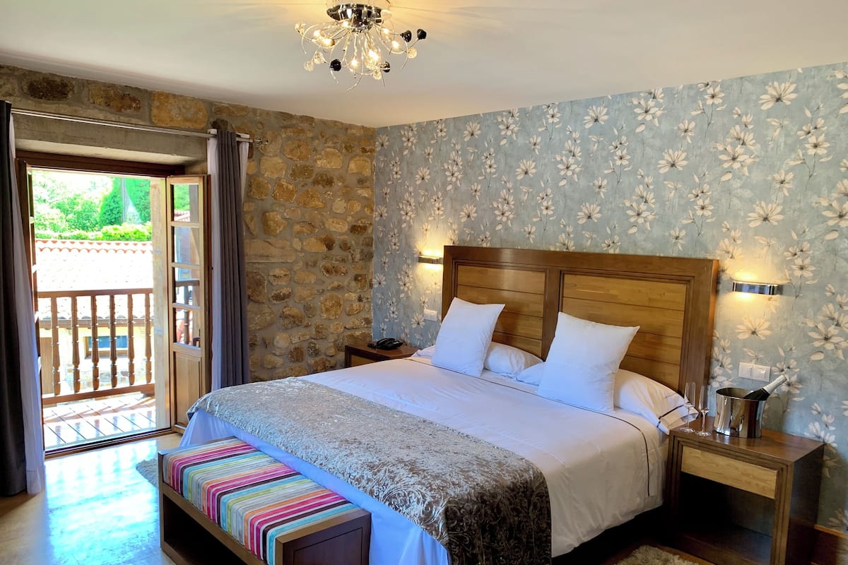 Een slaapkamer met een stenen muur, een houten hoofdbord, een bed met witte beddengoed, een nachtkastje met een fles en glazen, en een bankje met een gestreept kleed.