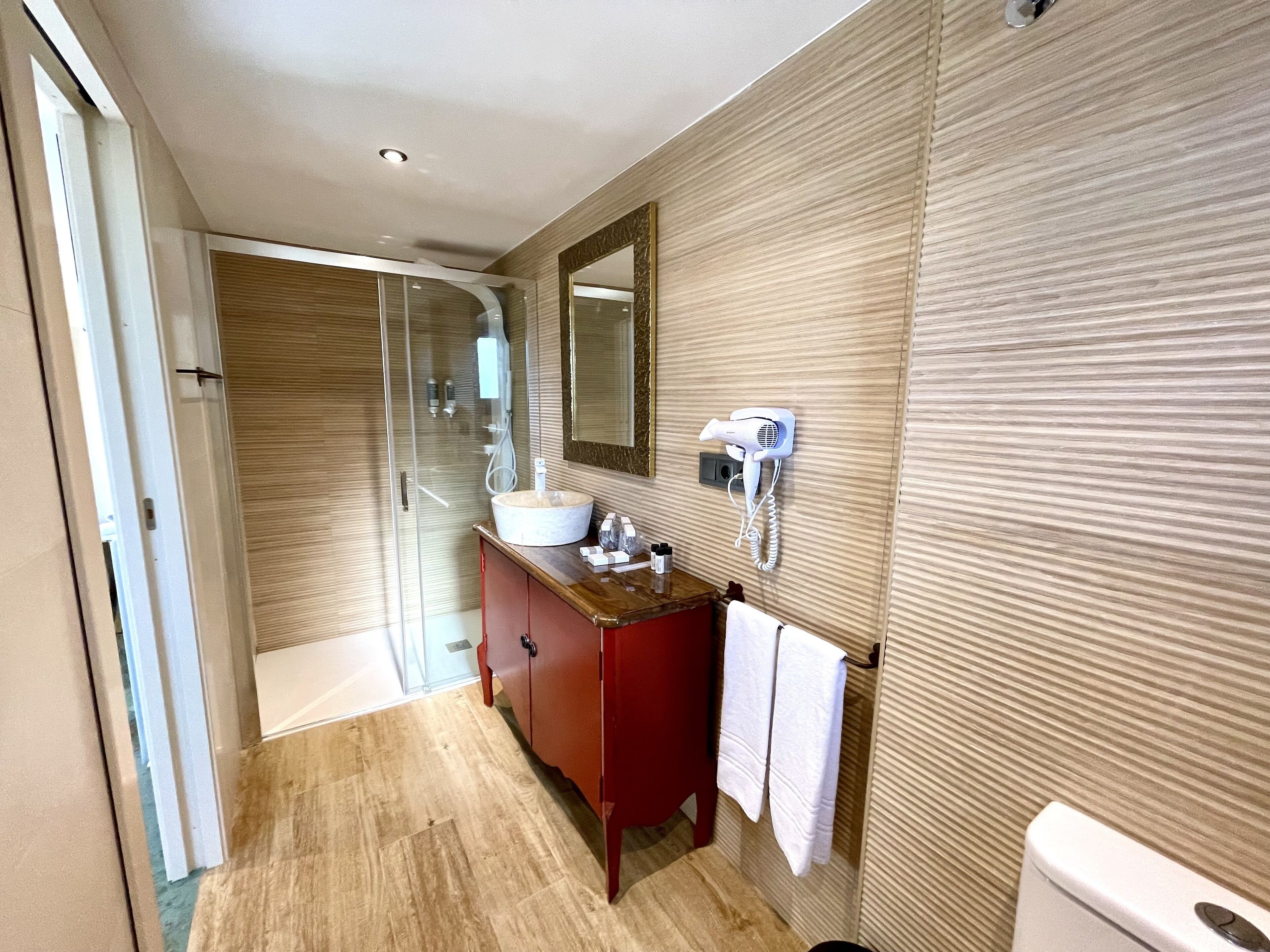 Un baño moderno con cabina de ducha de vidrio, tocador de madera, espejo grande y secador de pelo montado en la pared.
