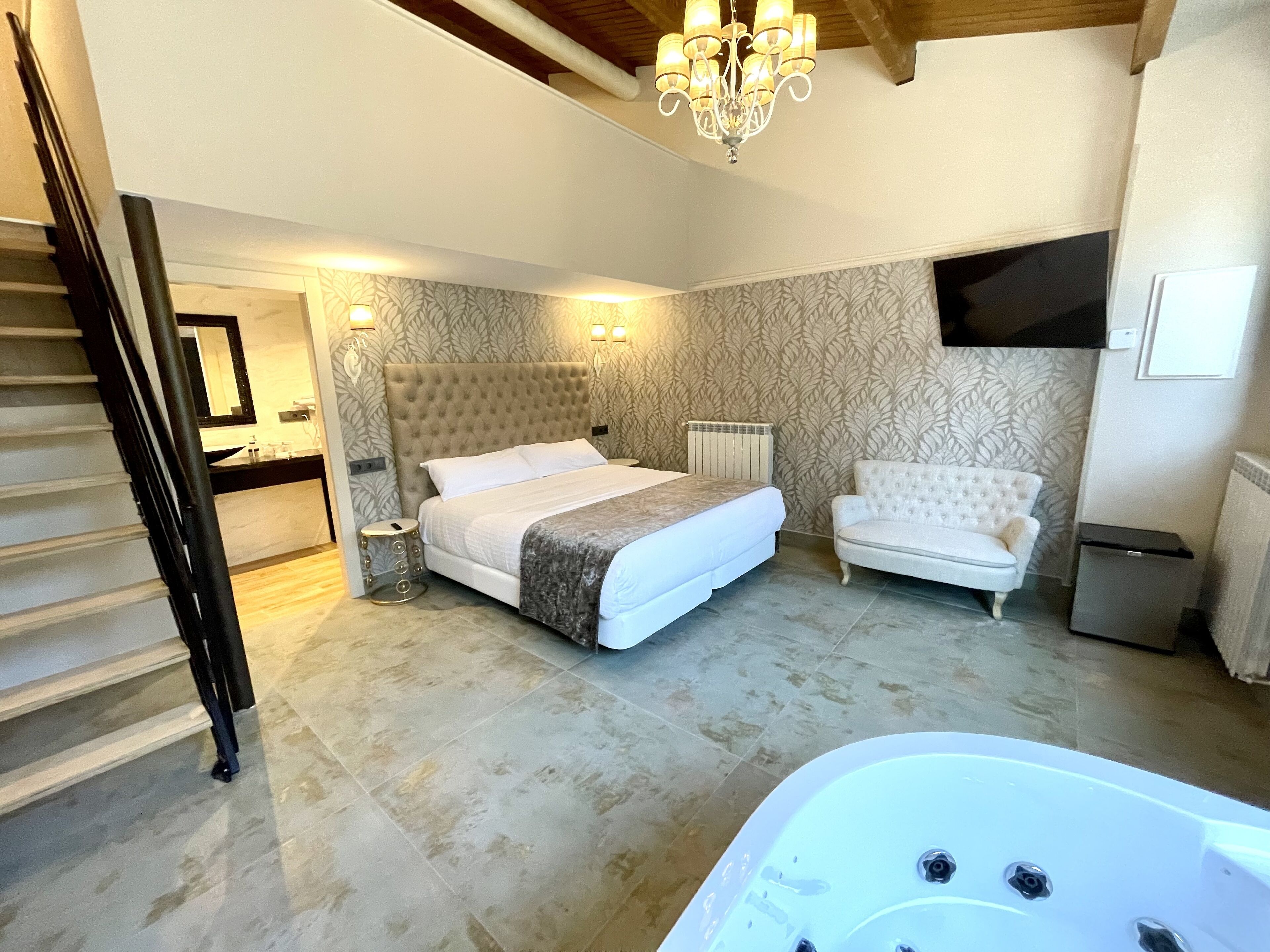 A bedroom with a large bed, a white sofa, a bathtub, and a chandelier.