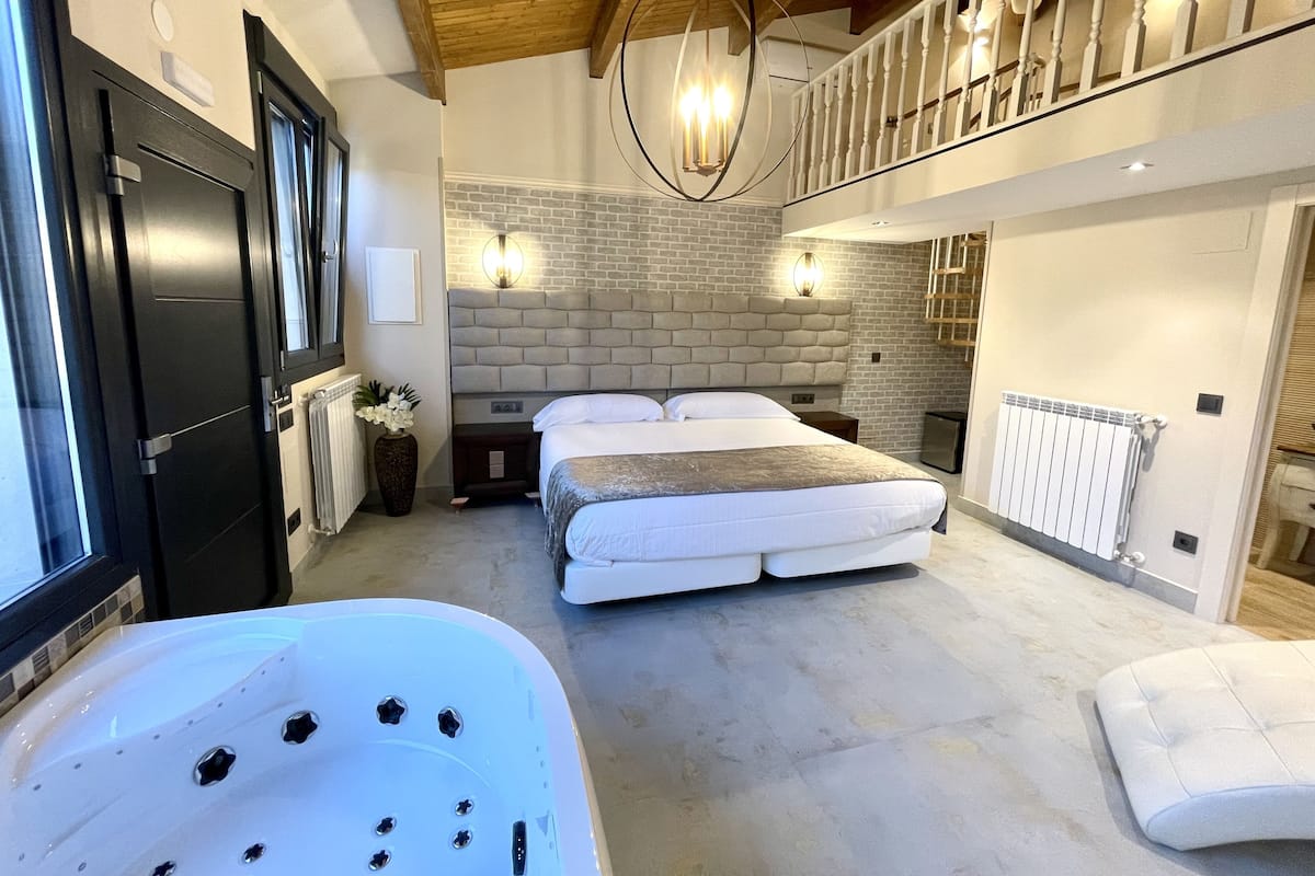 Een slaapkamer met een groot bed, een zithoek met een witte bank, een badkuip en een trap naar een hoger gelegen verdieping.