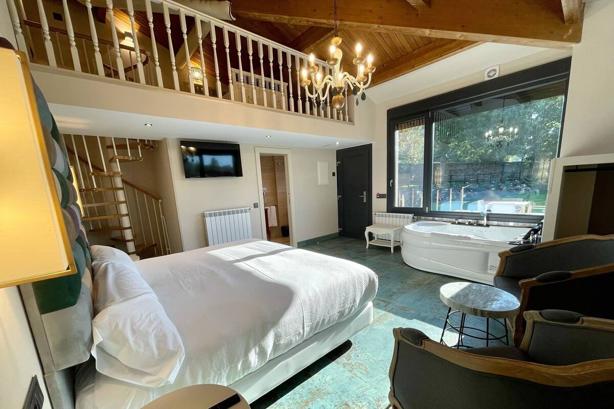 Hotelkamer met een bed, zithoek met een stoel en een klein tafeltje, groot raam met uitzicht op groen, badkuip en een trap naar een hoger gelegen niveau.