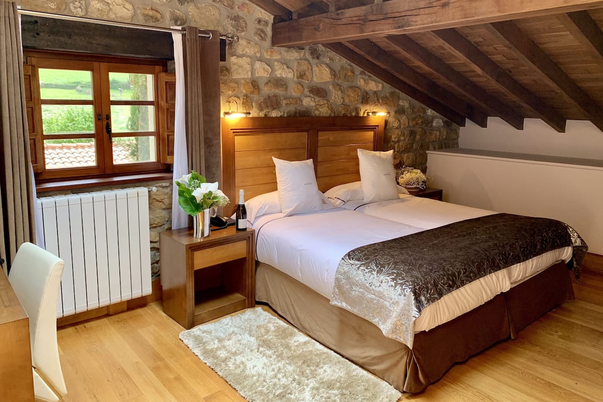 Een slaapkamer met een stenen muur, houten balken in het plafond, een bed met witte lakens, een nachtkastje met een bloevas en een raam met gordijnen.