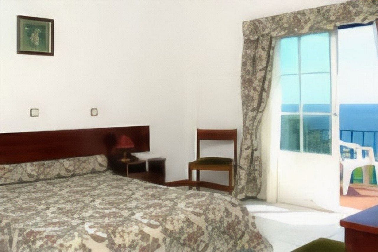 Habitación de hotel con cama, una silla, una mesita y ventana con vista al mar.