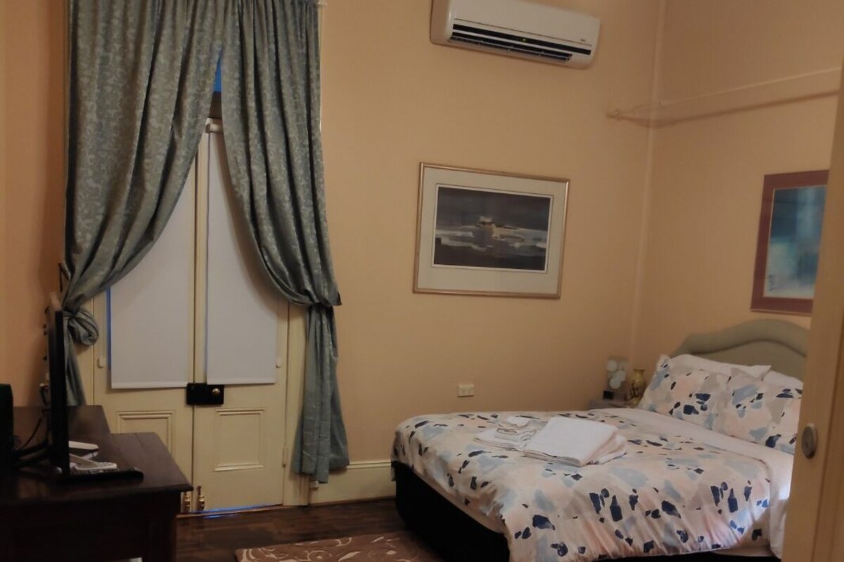 Ein Schlafzimmer mit einem Bett, einem Schreibtisch, einem Stuhl, einer Klimaanlage und einem Fenster mit Vorhängen.