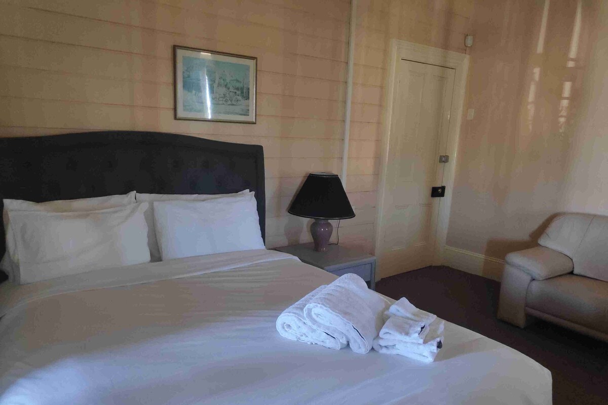 Ein Hotelzimmer mit einem großen Bett, einem Nachttisch mit Lampe, einem Sessel und einem gerahmten Bild an der Wand.