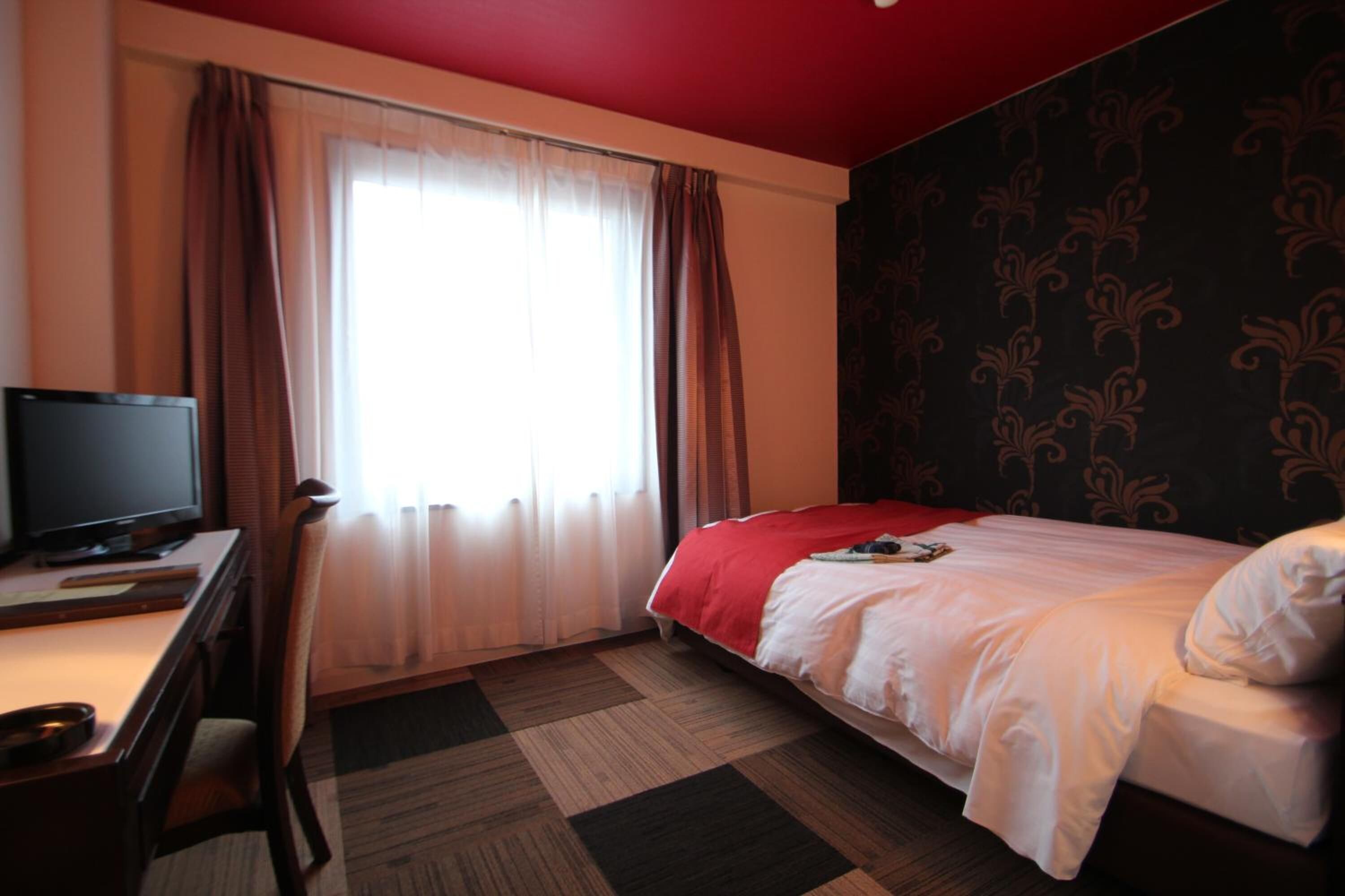 A hotel room with a bed, a desk with a television, and a window with curtains.