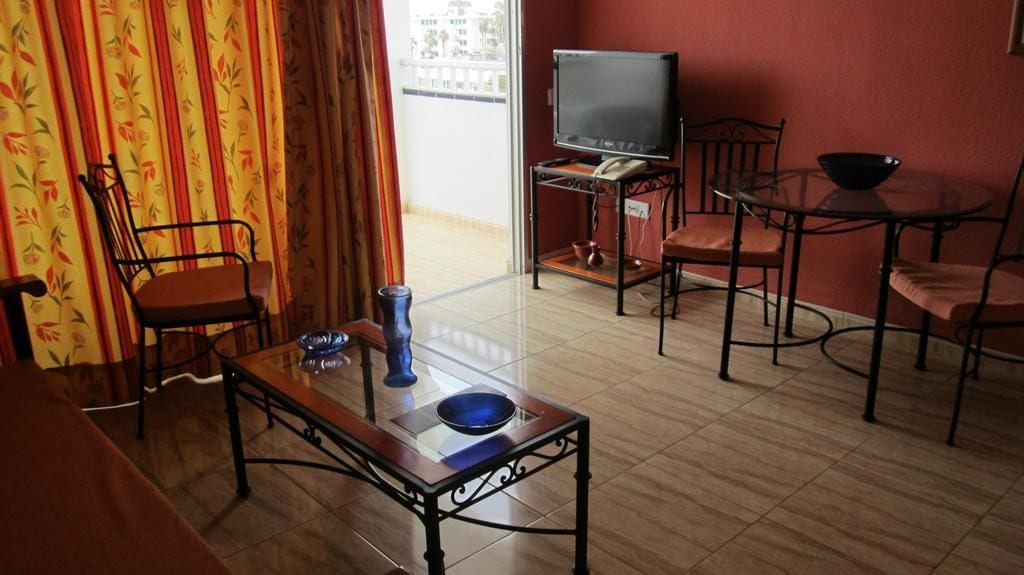 A living room with a television, a dining table with chairs, a sofa, and a window with curtains.