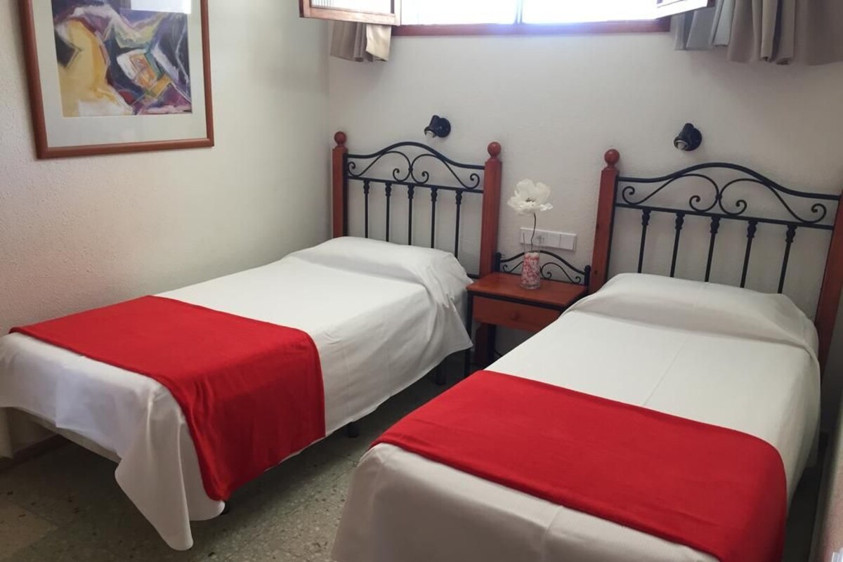 Two single beds with red and white bedding, a small bedside table with a vase, and a wall-mounted light fixture.