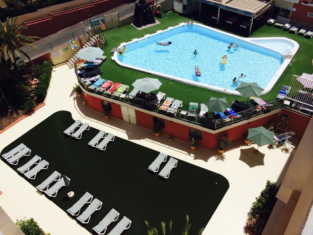 A swimming pool surrounded by lounge chairs and umbrellas, with a poolside area and a building in the background.