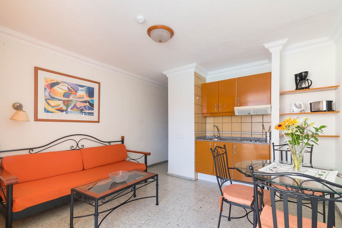 A living room with a sofa, coffee table, and chairs, a dining table with a vase of flowers, and a kitchen with orange cabinets and a stove.