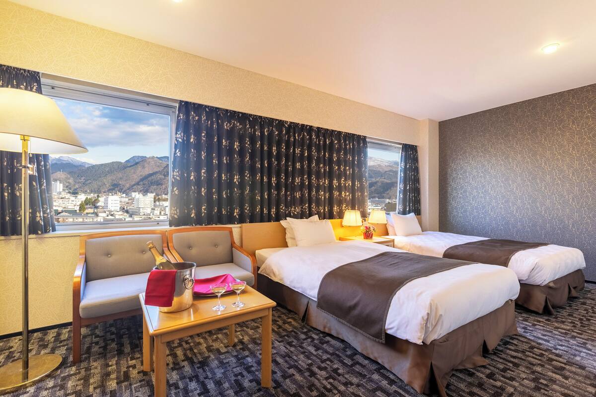 Een hotelkamer met twee bedden, een stoel, een tafel met een fles en glazen, en een uitzicht op bergen door het raam.