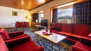 Habitación de hotel con sofá rojo, una mesita con una botella y vasos, televisor y vistas a las montañas a través de una ventana.