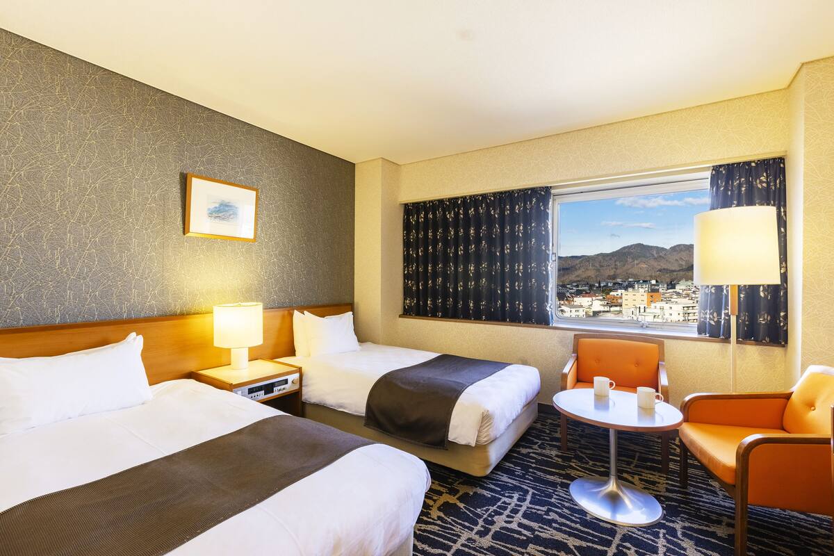 Een hotelkamer met twee bedden, een tafeltje, een stoel en een raam met uitzicht op een stadsgezicht.