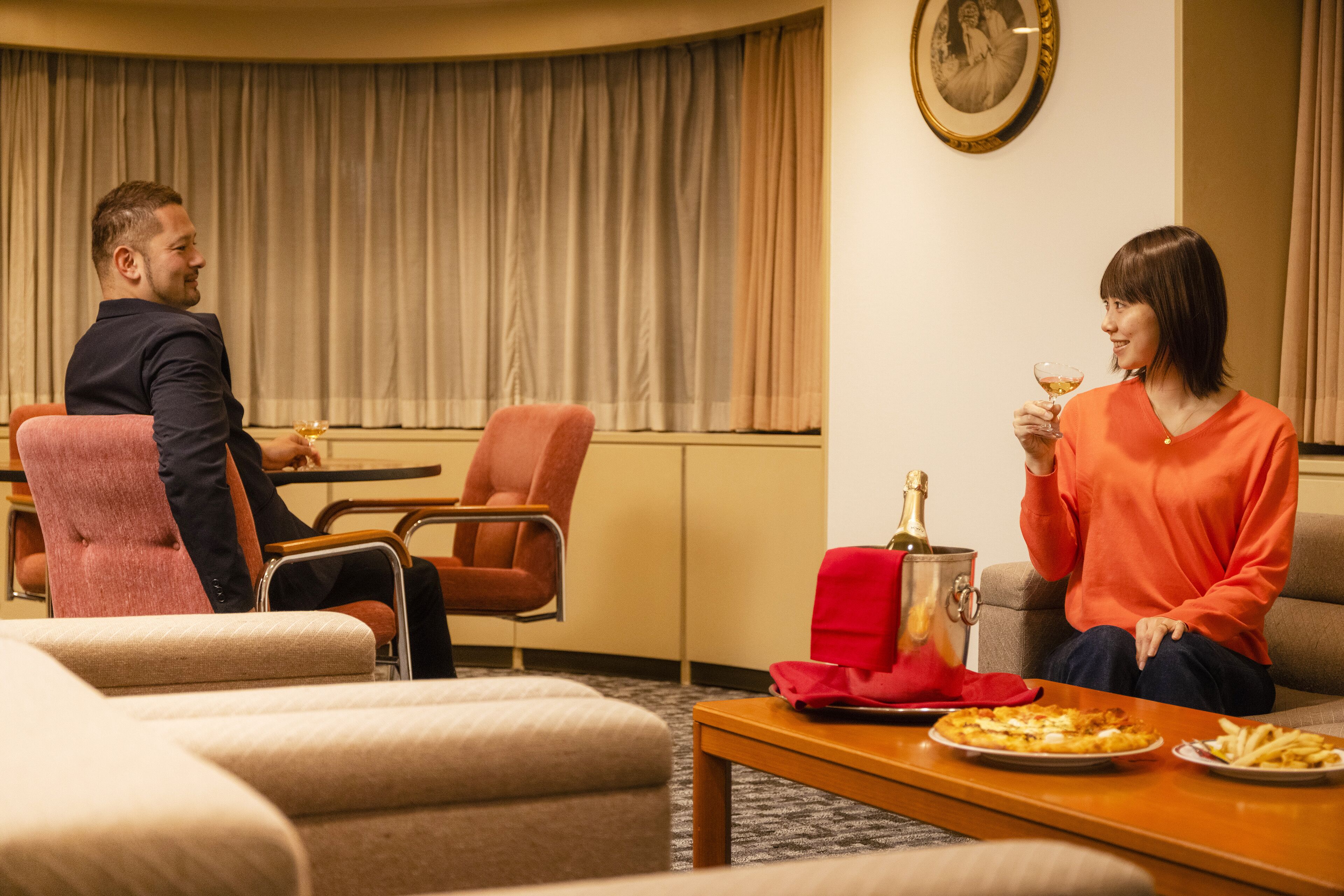 Een hotelkamer met een eettafel, twee stoelen, een bank en een groot raam met gordijnen.