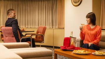 Een hotelkamer met een eettafel, twee stoelen, een bank en een groot raam met gordijnen.