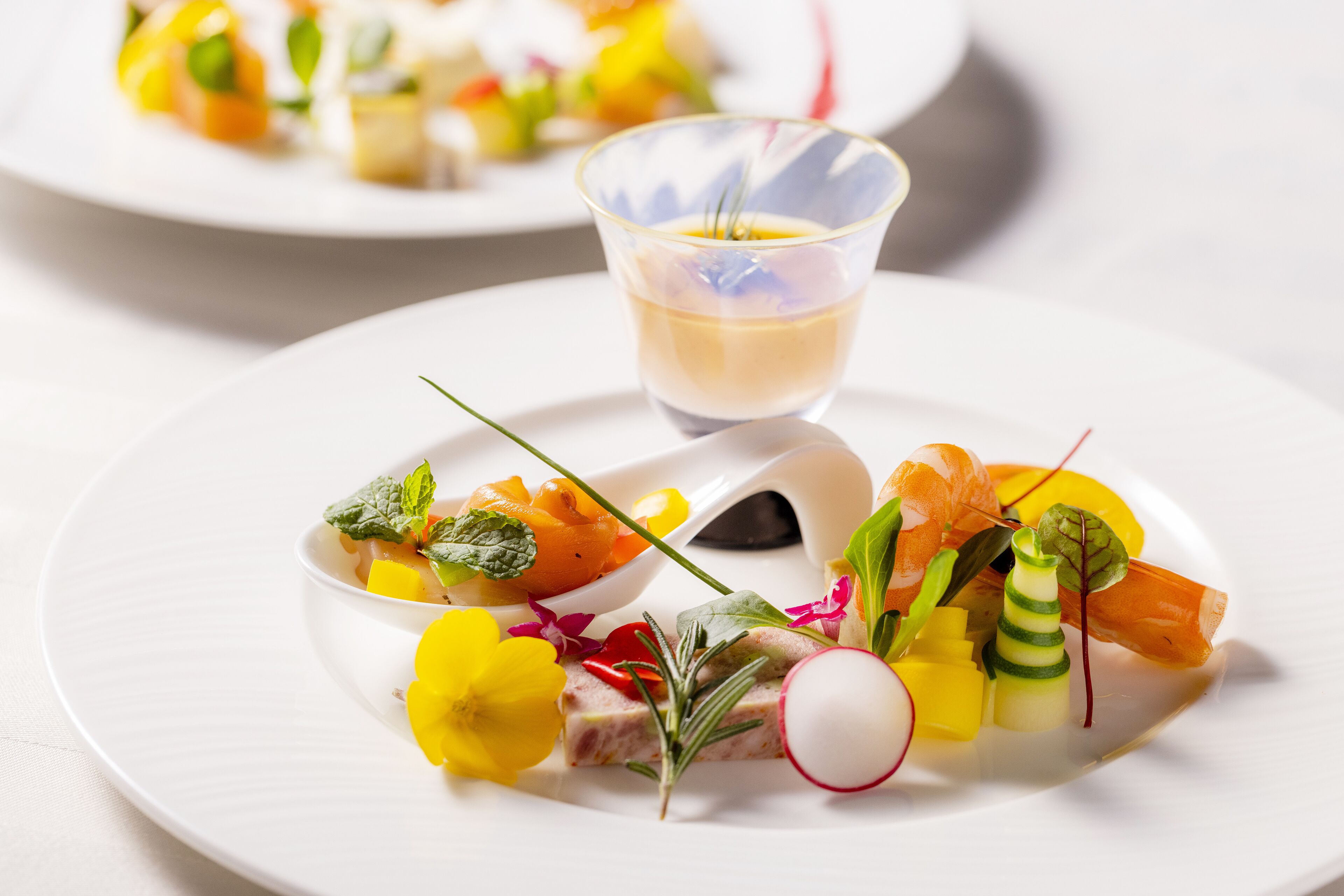 Une assiette blanche contenant une variété de légumes colorés et un petit verre d’un liquide jaune.