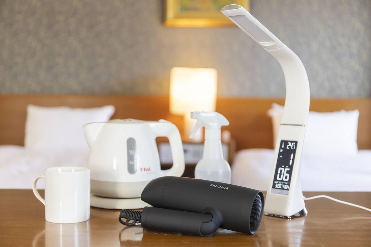Een hotelkamer met een bed, een witte mok, een zwarte draagbare speaker, een witte waterkoker en een digitale klok.