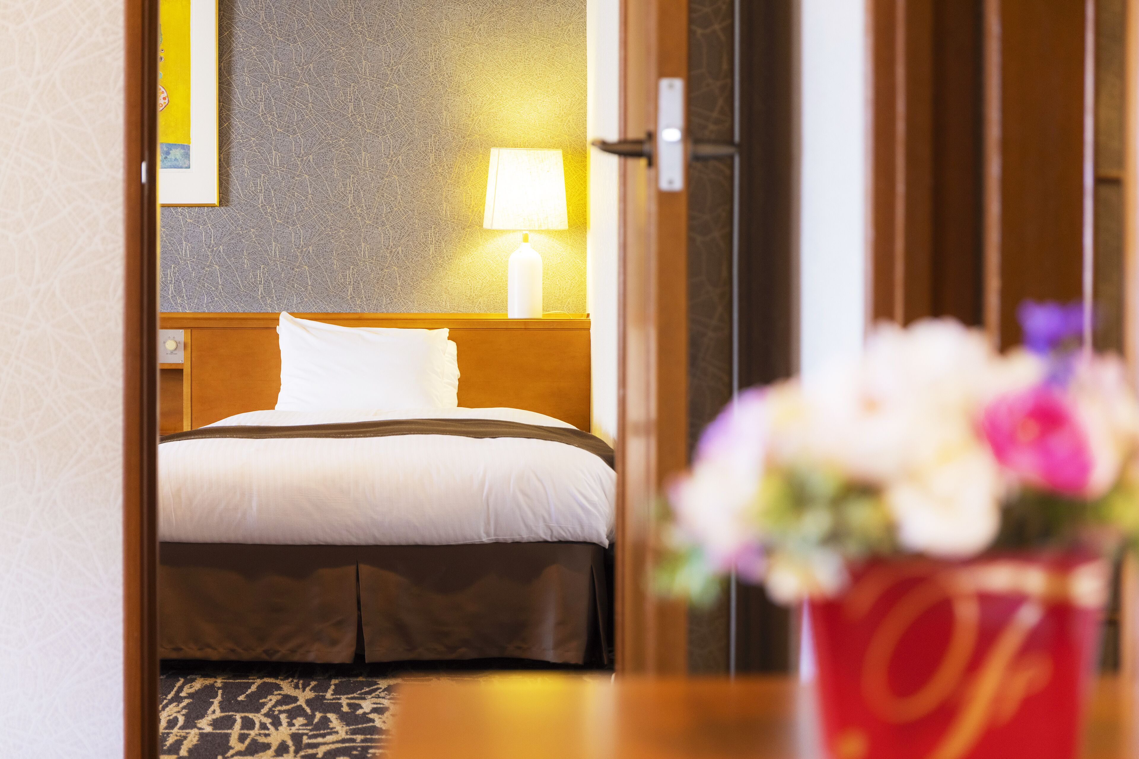 Une chambre d’hôtel avec un lit, une table de chevet avec une lampe et un arrangement floral sur la table.