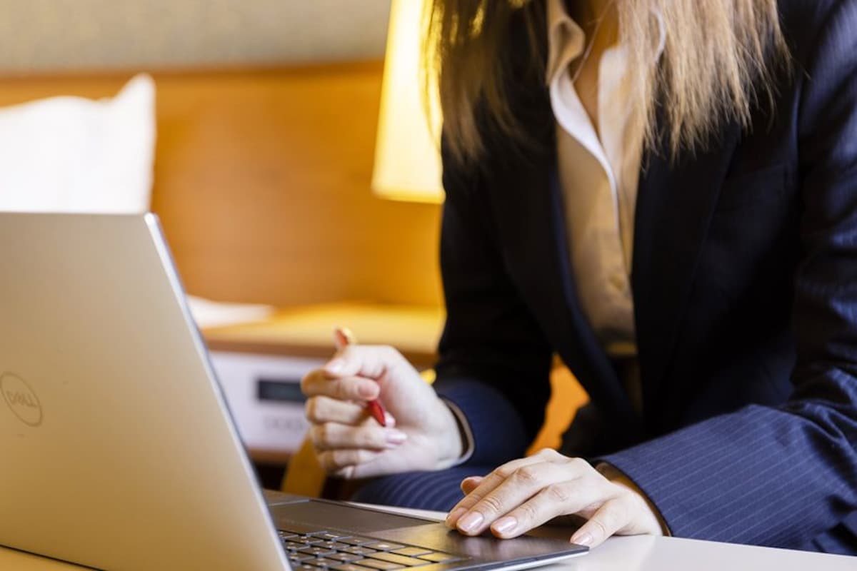 Una persona usando una computadora portátil sobre una mesa en una habitación con una cama y una lámpara al fondo.