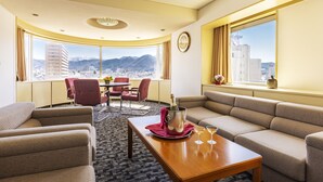 Habitación de hotel con un amplio ventanal con vista a la ciudad, un conjunto de sofás, una mesa de comedor con una botella y vasos, y una silla roja.