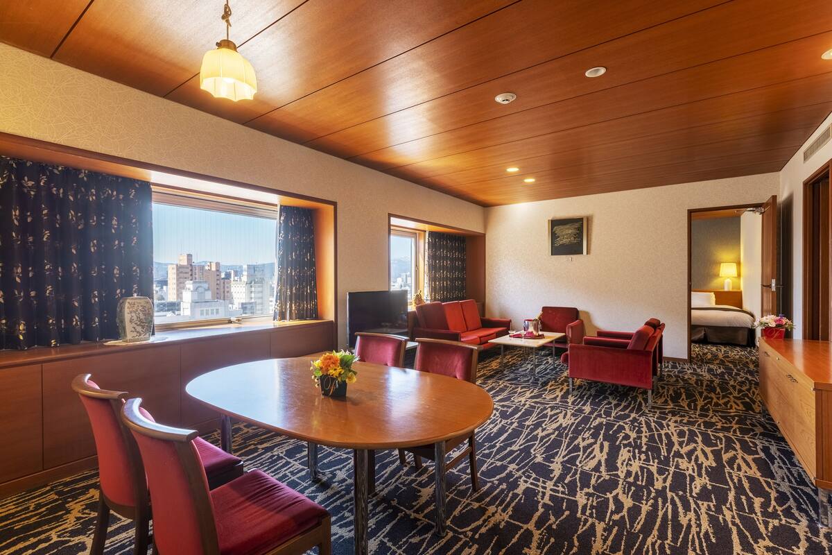 Een hotelkamer met een groot raam, een ronde tafel met stoelen, een bank en een kleine televisie.
