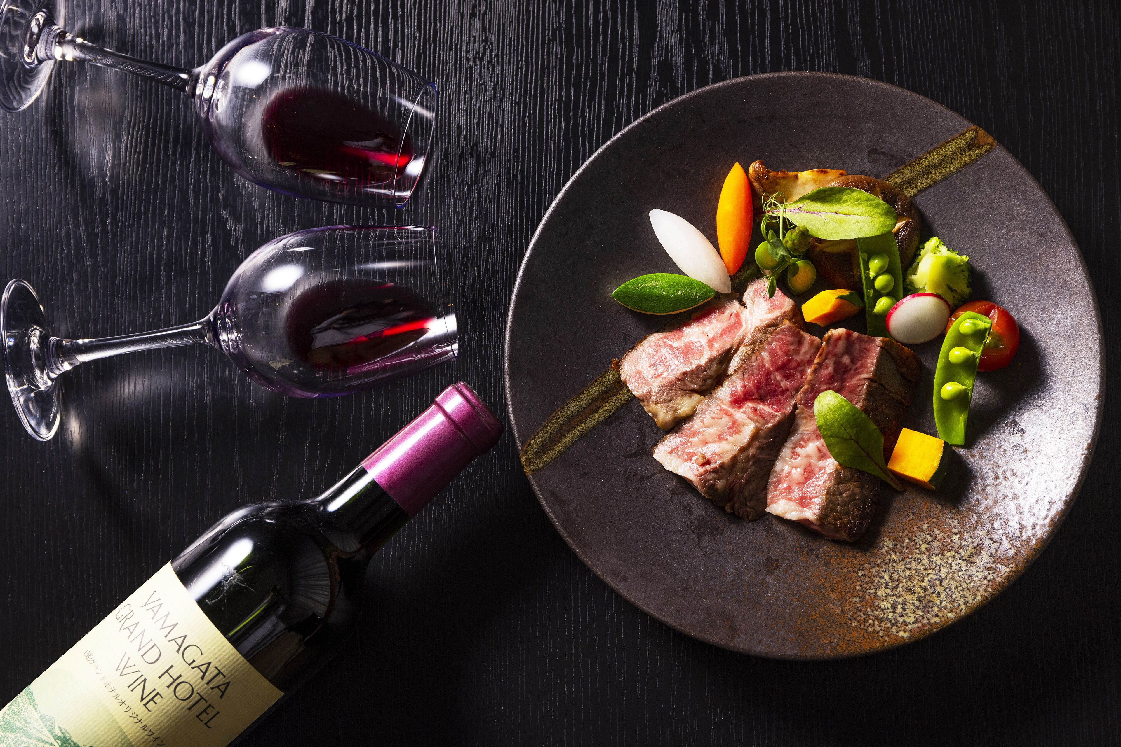 Eine Flasche Wein vom Yamagata Grand Hotel, ein Teller mit Steak und Gemüse sowie zwei Weingläser auf einem dunklen Holztisch.
