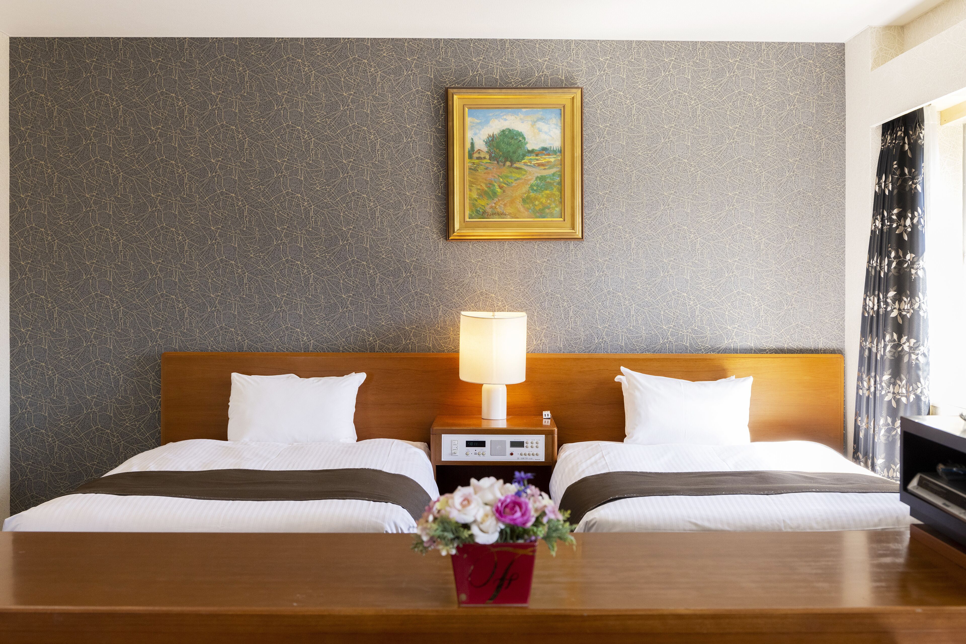 Habitación de hotel con dos camas, una mesita de noche con lámpara, un cuadro en la pared y un florero en la mesa.