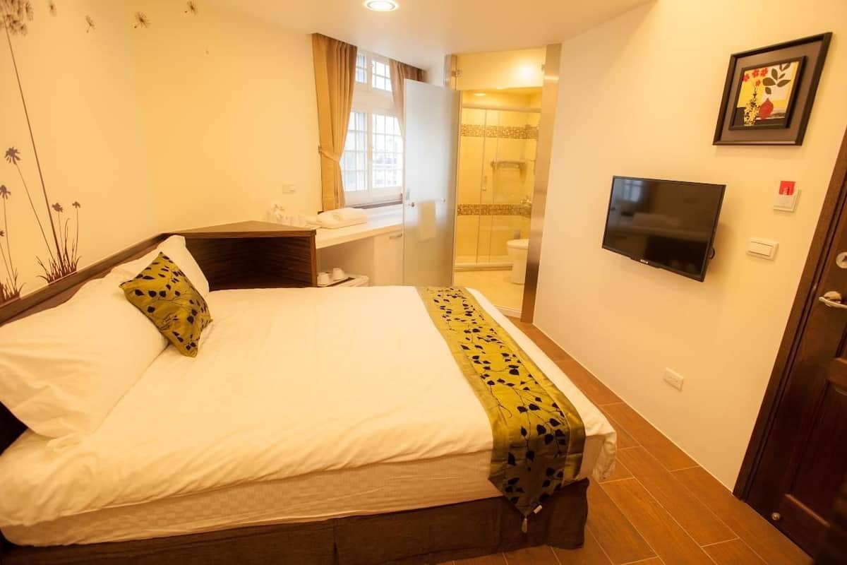 A hotel room with a bed, a TV, a bathroom with a shower, and a window with curtains.
