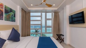 Una habitación de hotel con una cama, ventilador de techo, televisor, ventana con vista al océano y obras de arte en la pared.