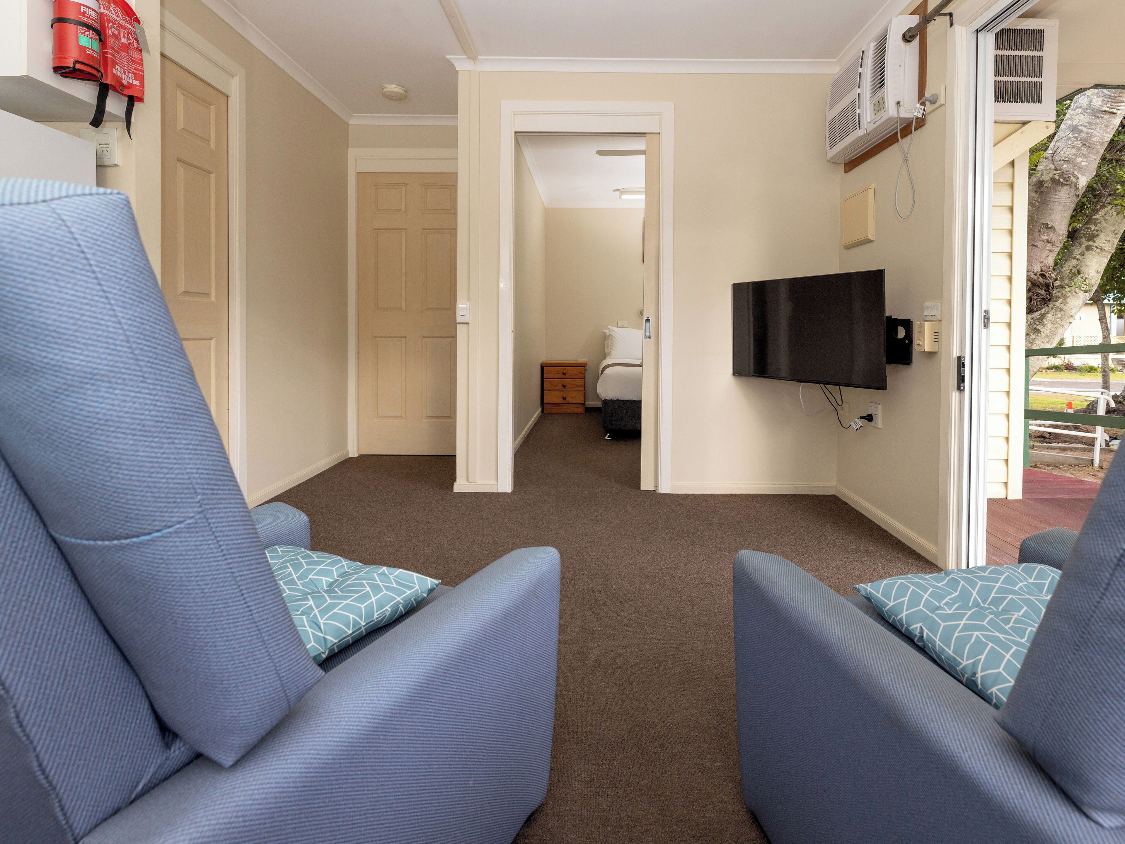 A hotel room with a large window, a flat-screen TV mounted on the wall, and a door leading to a bedroom.