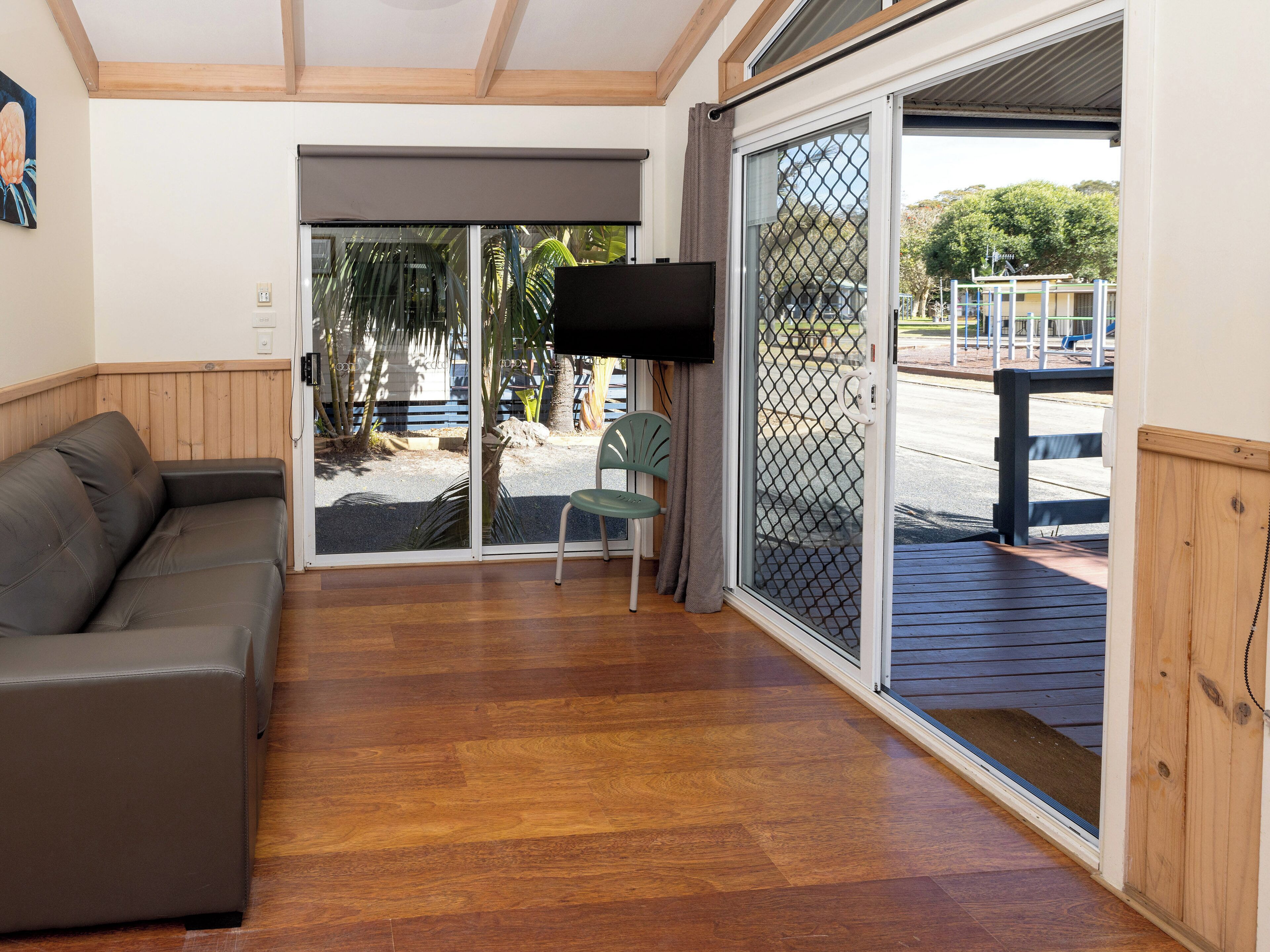 A room with a wooden floor, a grey sofa, a green chair, a TV, and a sliding glass door leading to an outdoor area with a playground.