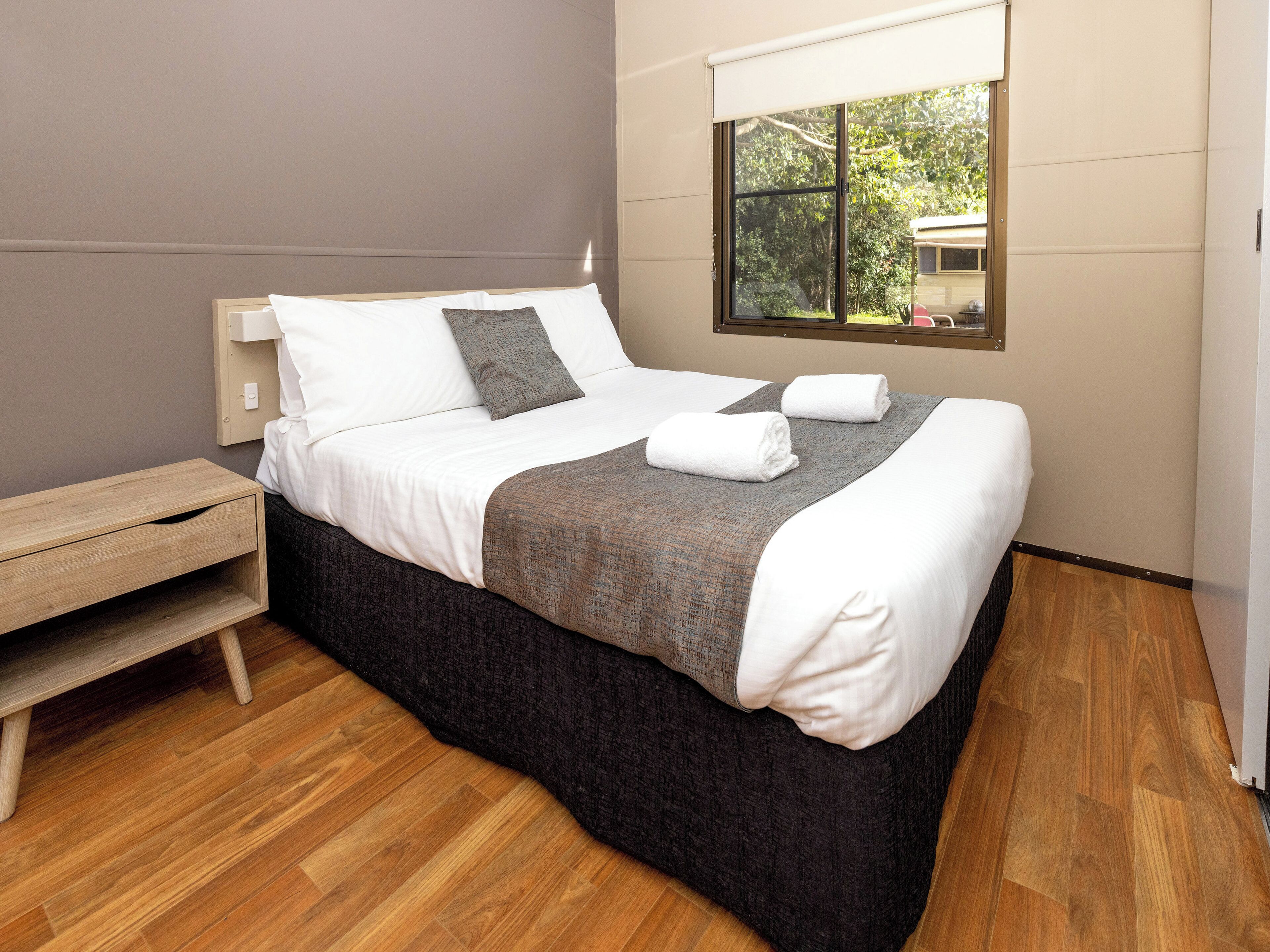 Een net opgemaakt bed met witte en grijze lakens, een houten nachtkastje en een raam met uitzicht op groen.