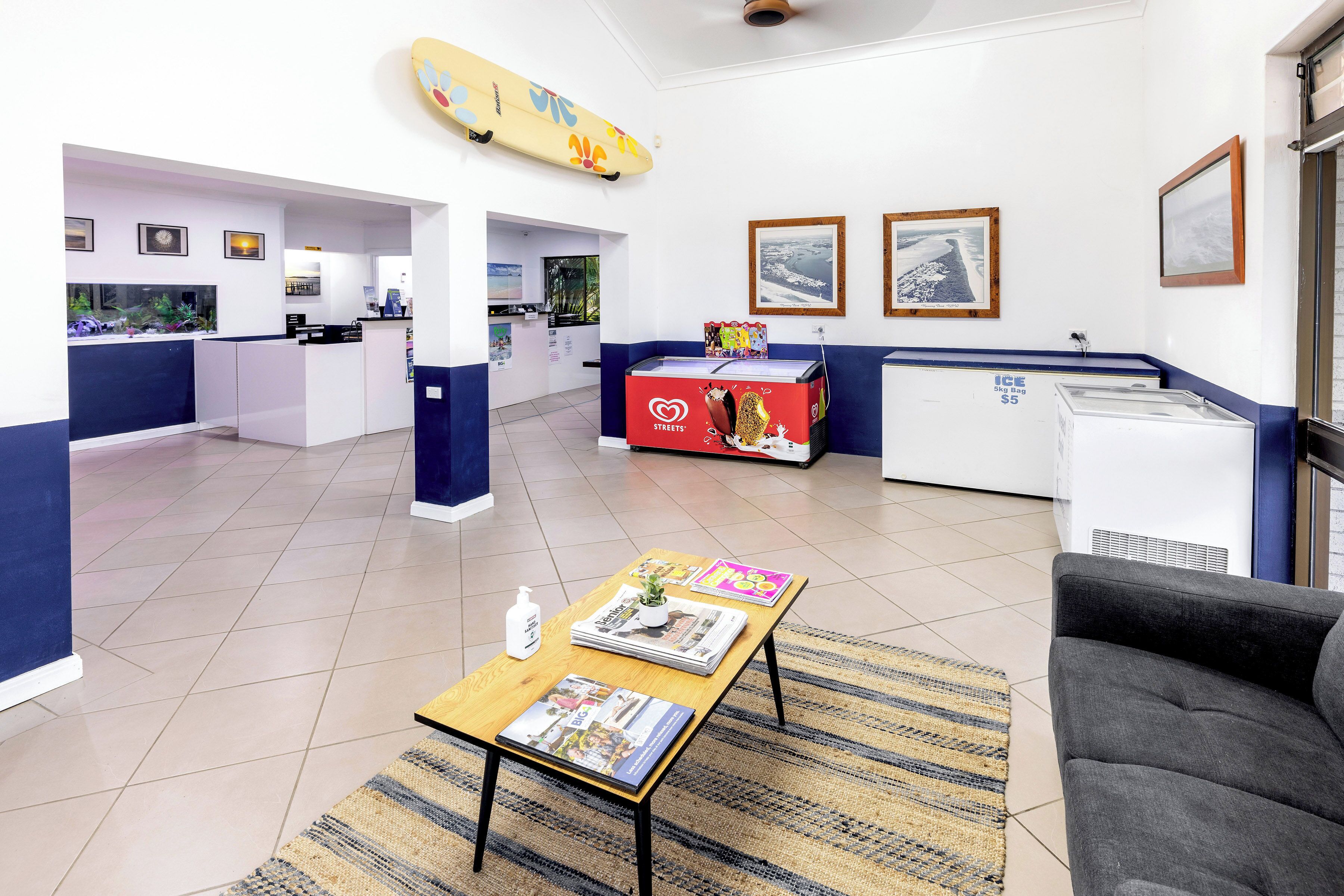Un moderno vestíbulo de hotel con mostrador de recepción, zona de estar con revistas, decoración de tabla de surf y refrigerador.