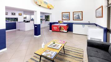 Un moderno vestíbulo de hotel con mostrador de recepción, zona de estar con revistas, decoración de tabla de surf y refrigerador.