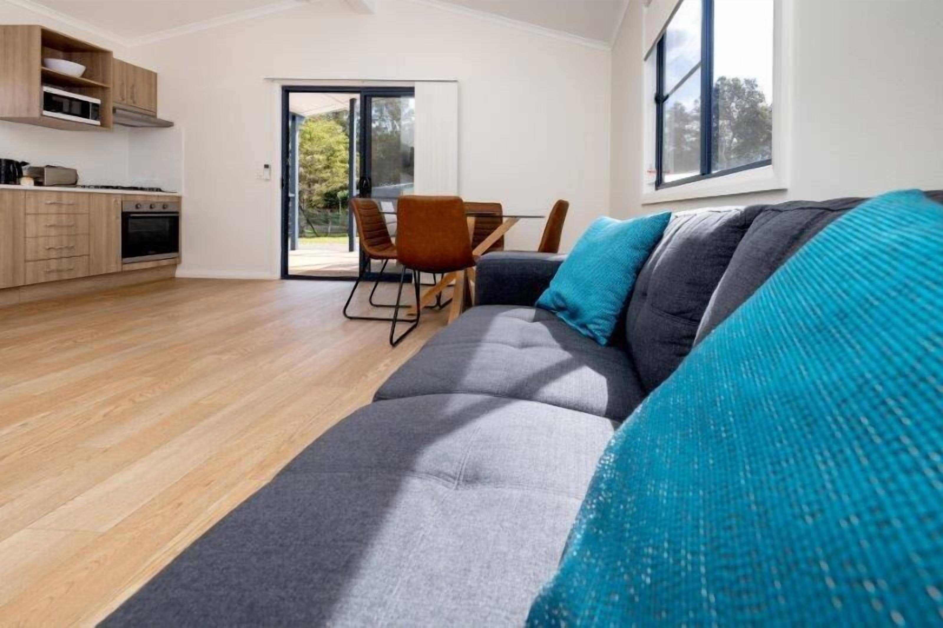 A modern living room with a grey sofa, a dining table with chairs, and a kitchen area with wooden cabinets and a stove.