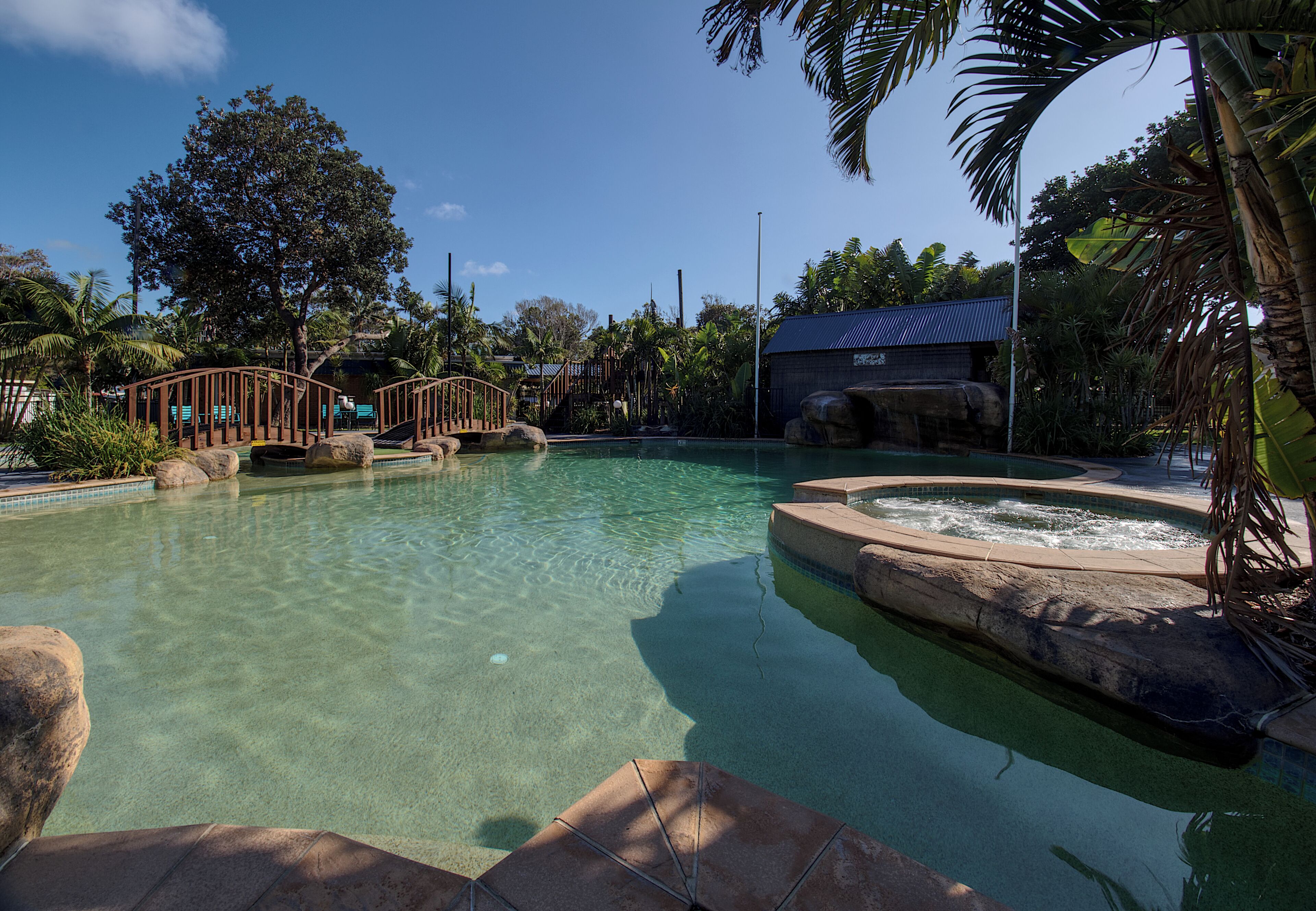 A clear pool with a wooden bridge, a hot tub, and tropical vegetation.