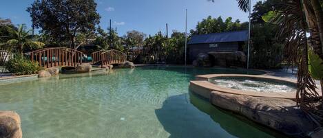A clear pool with a wooden bridge, a hot tub, and tropical vegetation.