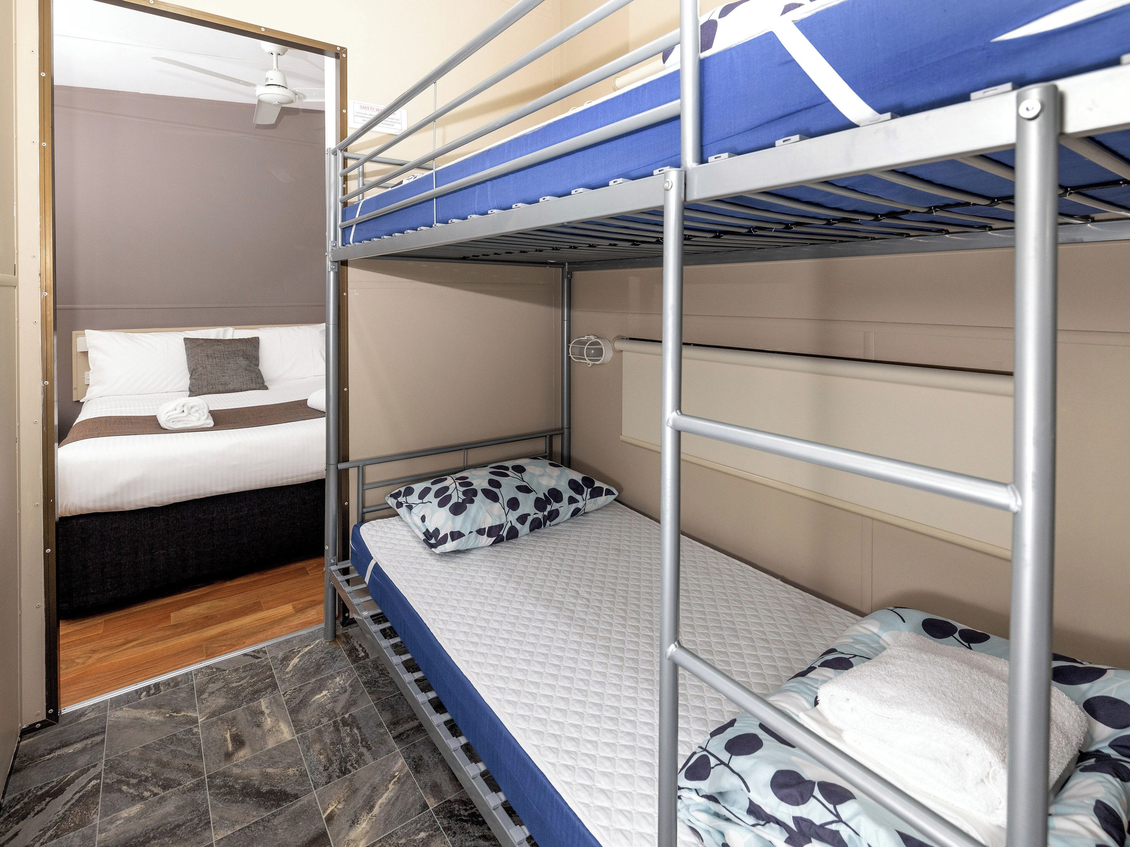 Ein Schlafraum mit Etagenbetten, einem Deckenventilator, Holzboden und einem Bett mit weißer Matratze sowie einem Kissen in blau-weißer Optik.
