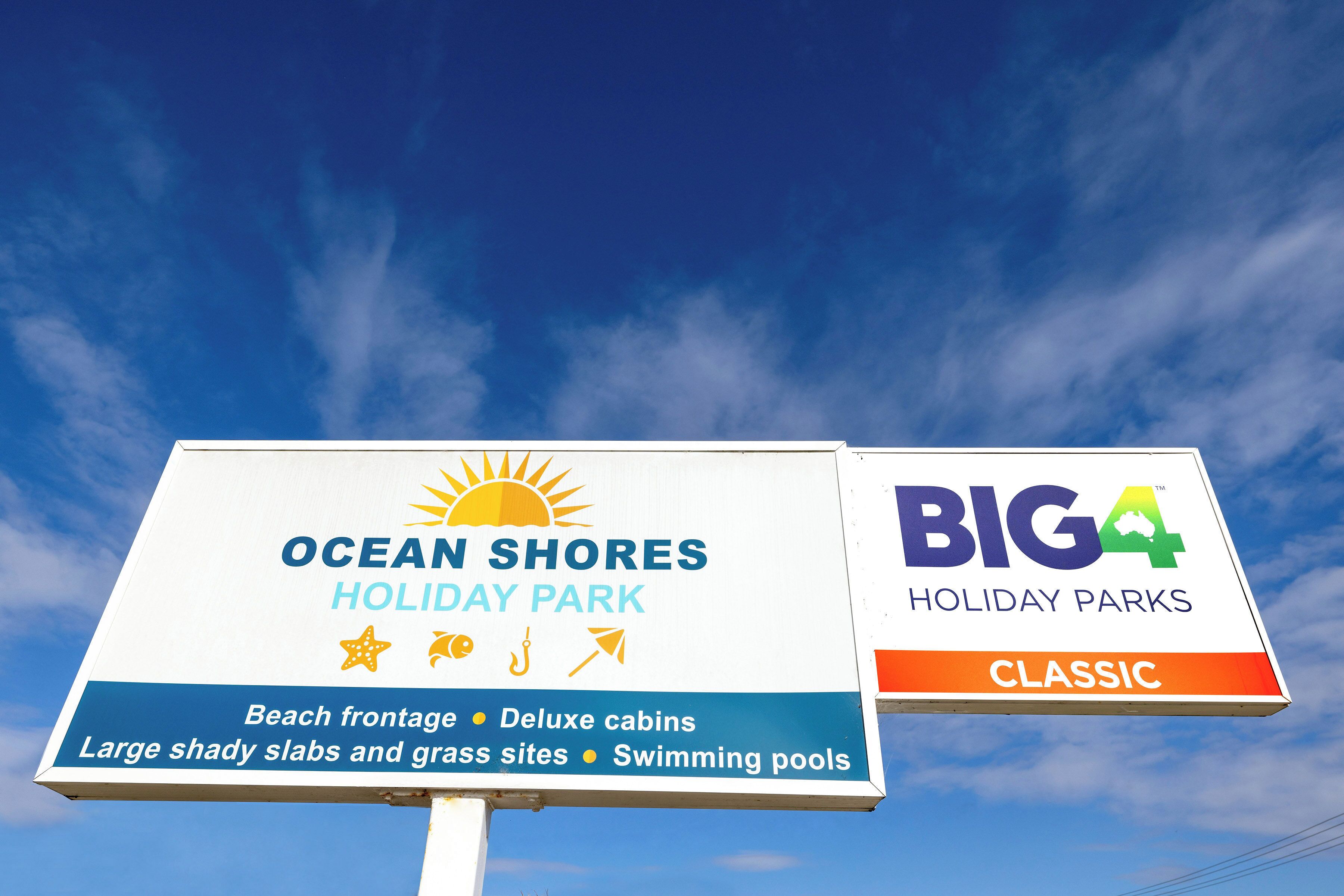 A sign for Ocean Shores Holiday Park, highlighting features like beach frontage, deluxe cabins, large shady slabs, and swimming pools.