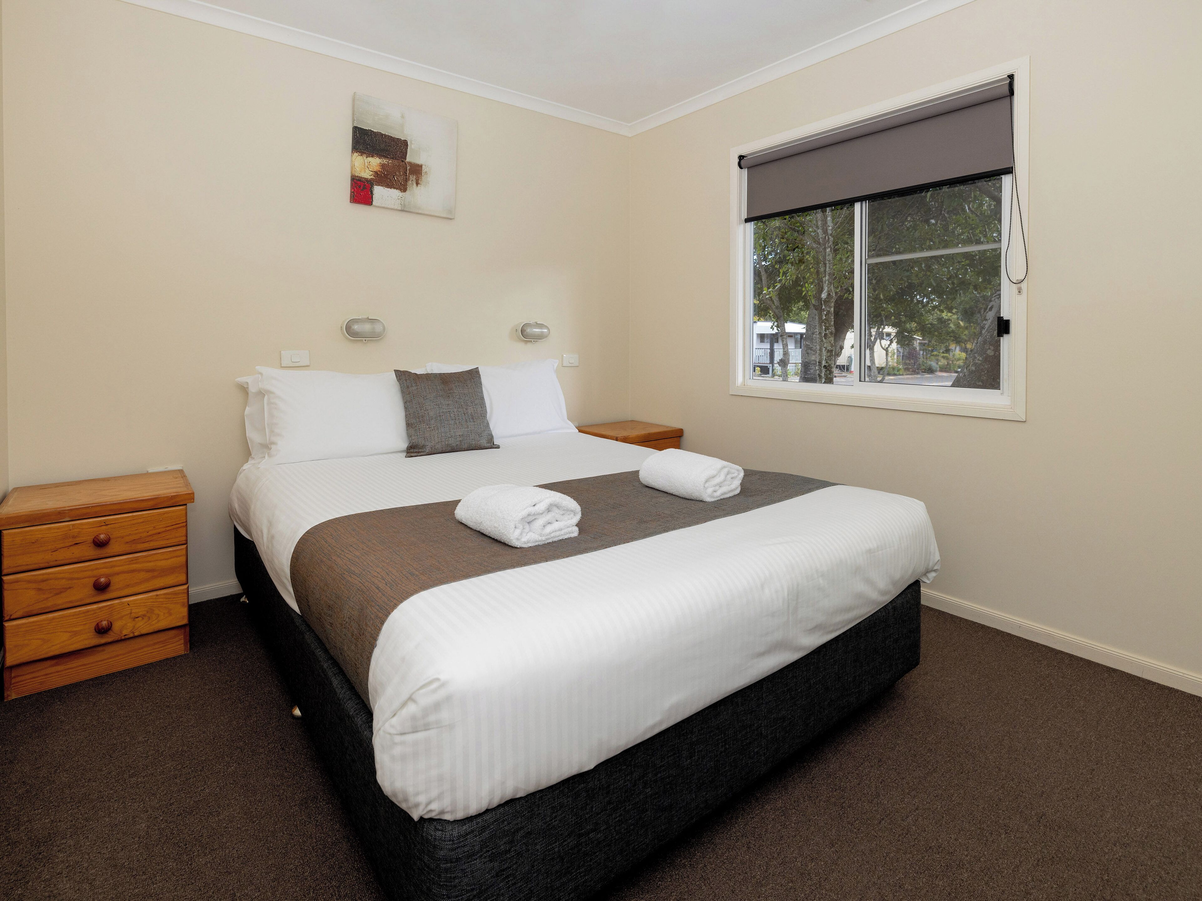 A neatly made bed with white linens and two folded towels, a wooden nightstand with drawers, a gray pillow, and a wall-mounted artwork.