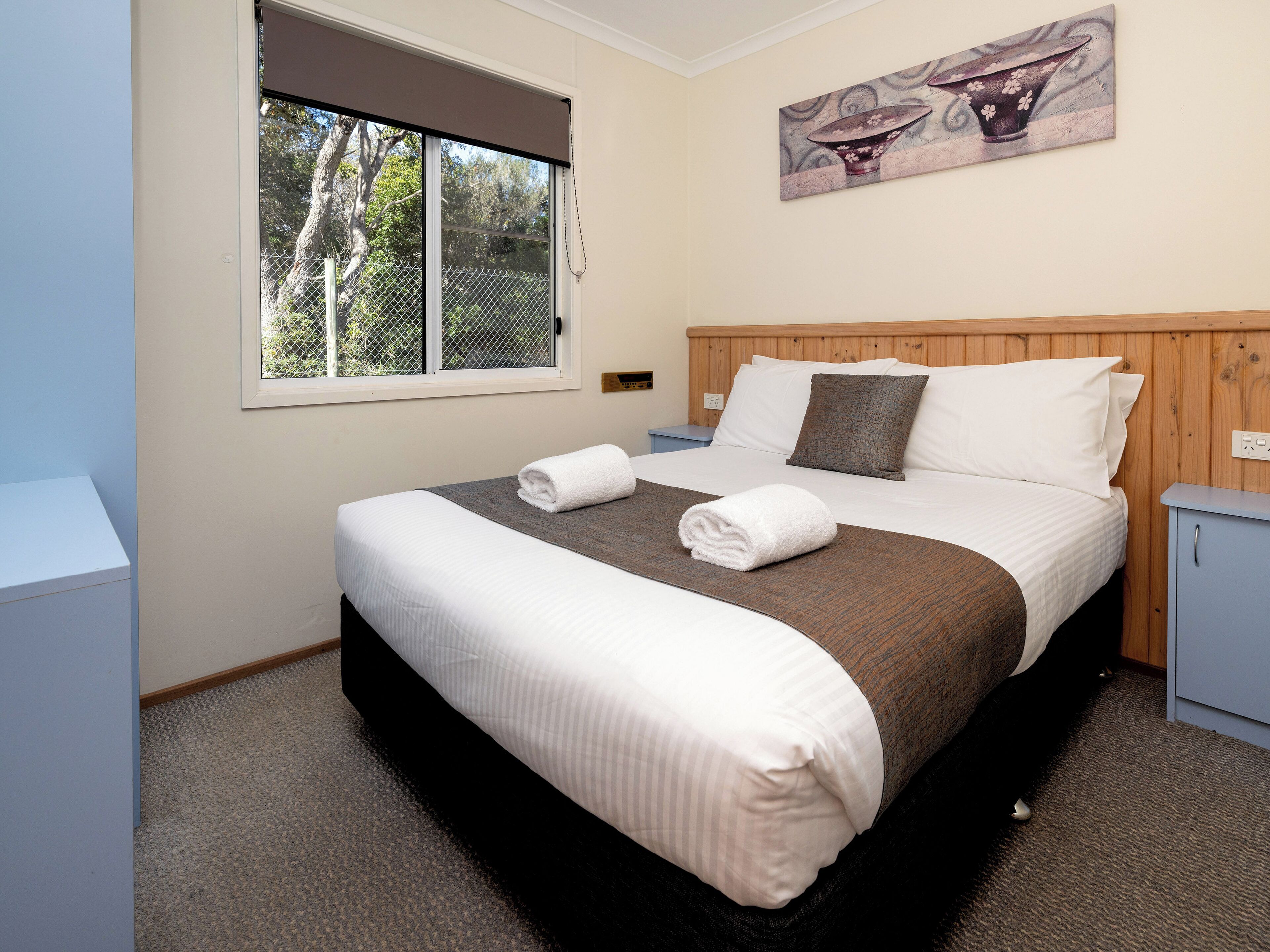 A neatly made bed with white and brown bedding, a wooden headboard, and a painting above it.